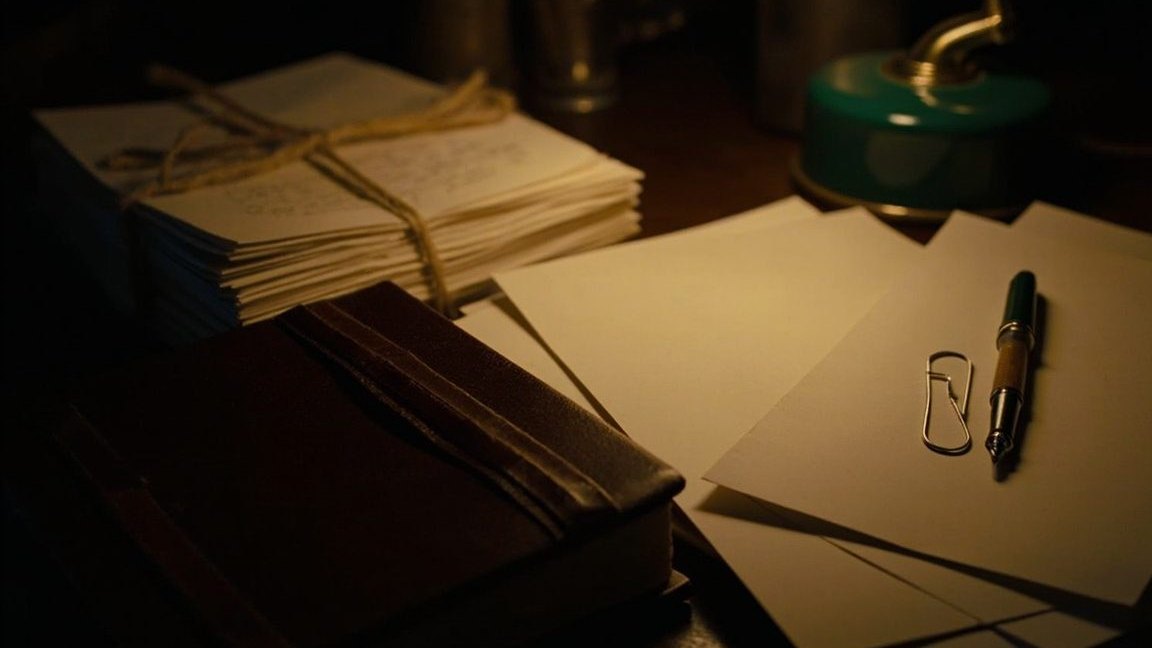 A closed brown leather ledger on a dark wooden desk beside a bundle of paper slips tied with string, loose sheets, a paperclip and a fountain pen under a green desk lamp
